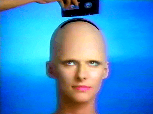 A hand slots a VHS-tape into the top of a bald womans head